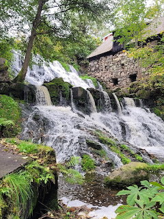 Cascading waterfall over mossy rocks with old stone building and green trees in background