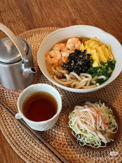 Korean udon bowl with shrimp, egg strips, and seaweed, served with tea and a small salad on a woven tray