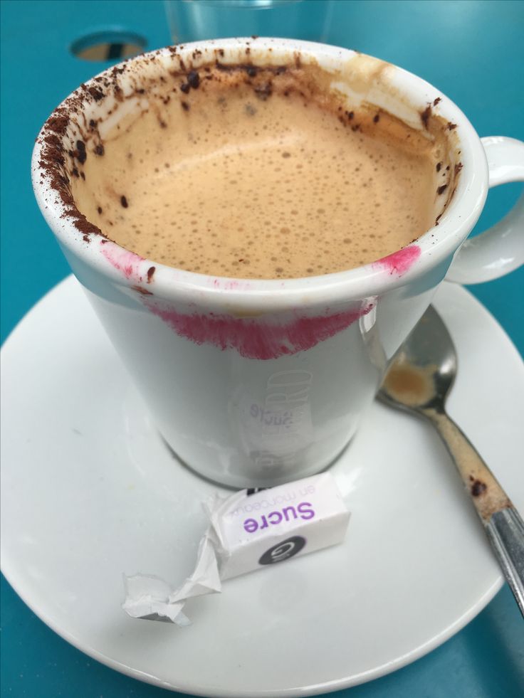 Coffee cup with foam and cocoa powder, pink lipstick stain visible on rim, white saucer with sugar packets