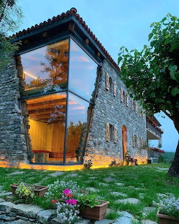 Modern stone house with illuminated angular windows at dusk, surrounded by garden and trees