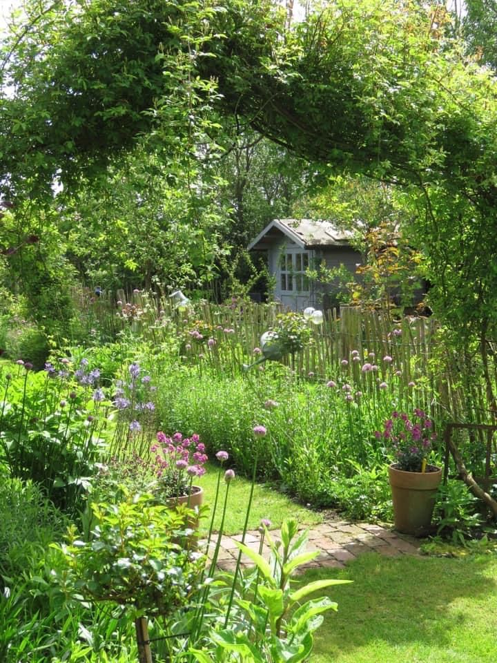 Flower garden with a curved path, wooden fence, and a small cottage under trees
