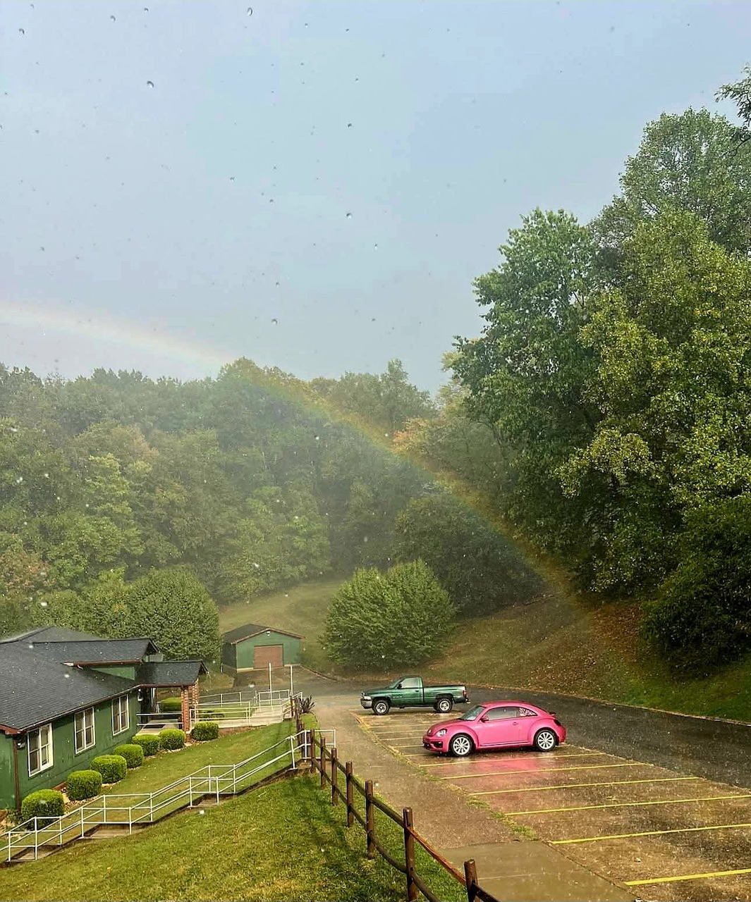 Pink and green cars in a rainy parking lot with a faint rainbow over wooded hills