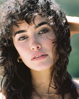 Close-up portrait of a young woman with dark curly hair, green eyes, and gold earing outdoors