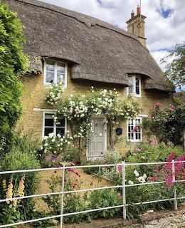 Thatched cottage with climbing roses, flowers, and a white fence in front