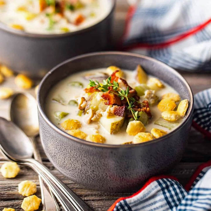 Bowl of creamy clam chowder topped with herbs and bacon, with crackers on a wooden table