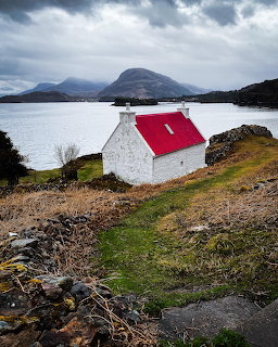 White cottage with red roof on a Scottish coastline with mountains and overcast sky
