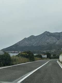 Road leading toward a large rocky mountain under grey overcast sky with town at its base