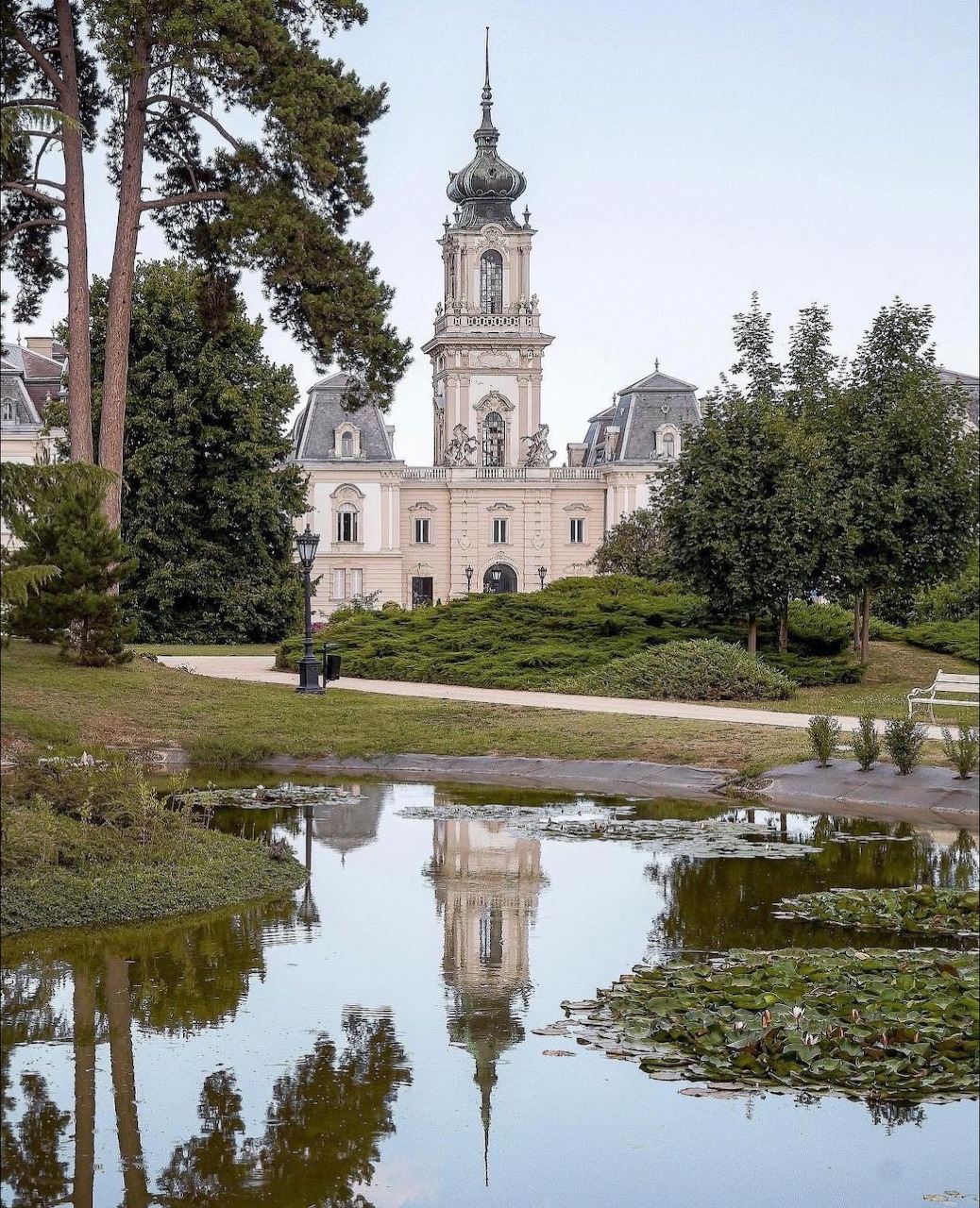 White baroque palace with clock tower reflected in pond, surrounded by trees and lawn