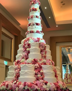 Tall white tiered cake covered with pink and white flowers in an indoor venue