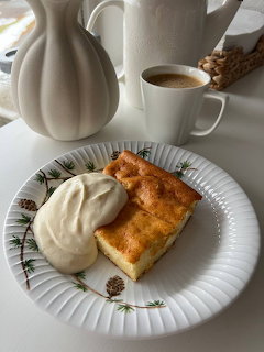 Slice of cake with cream on plate next to coffee cup and white vase
