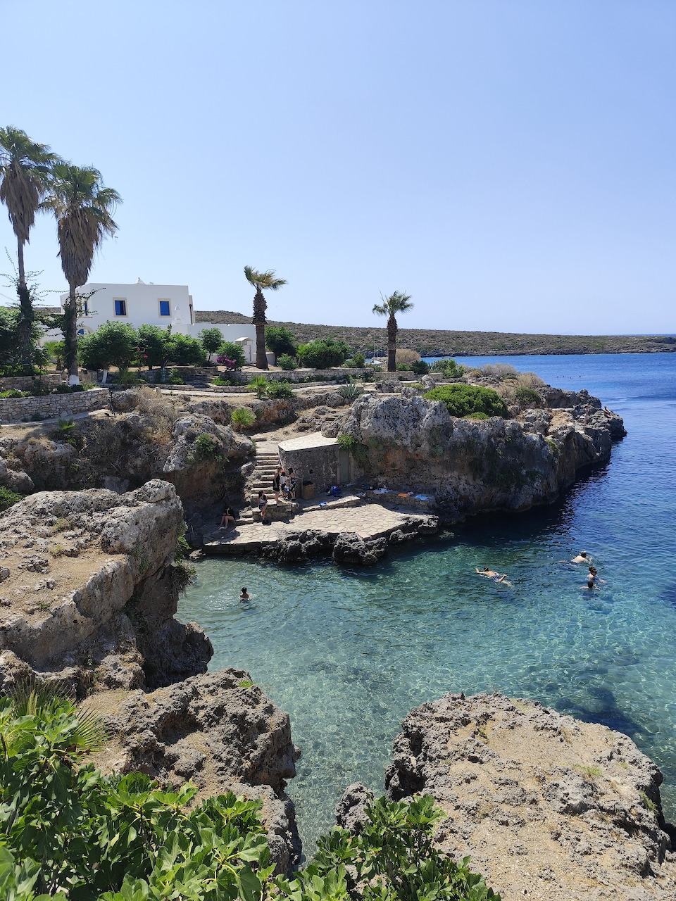 Rocky cove with turquoise water, palm trees, and a white building at Avlemonas, Kithira, Greece