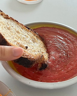 Hand dipping crispy toast into bowl of red tomato soup with olive oil drizzle and pepper