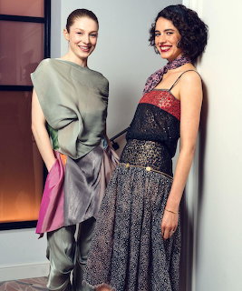 Two women posing together indoors wearing stylish outfits, smiling at the camera
