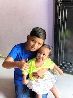 Smiling boy in blue shirt holding a baby girl in yellow top, posing together indoors