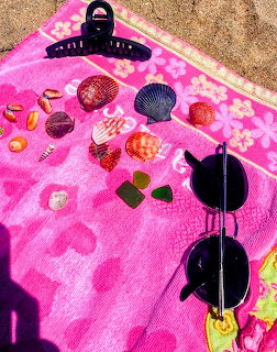Seashells, sea glass, sunglasses and a hair clip arranged on a pink beach towel on sand