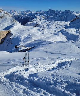 Skis and poles stuck in snow at a mountain summit overlooking snowy Alpine peaks under blue sky