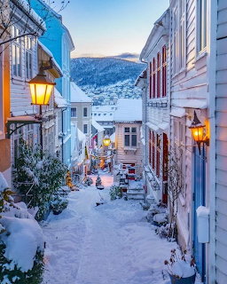 Snowy narrow street with wooden buildings, lit street lamps, and mountain backdrop at twilight