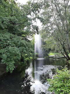 Tall water fountain shooting up from a pond surrounded by green trees in a park