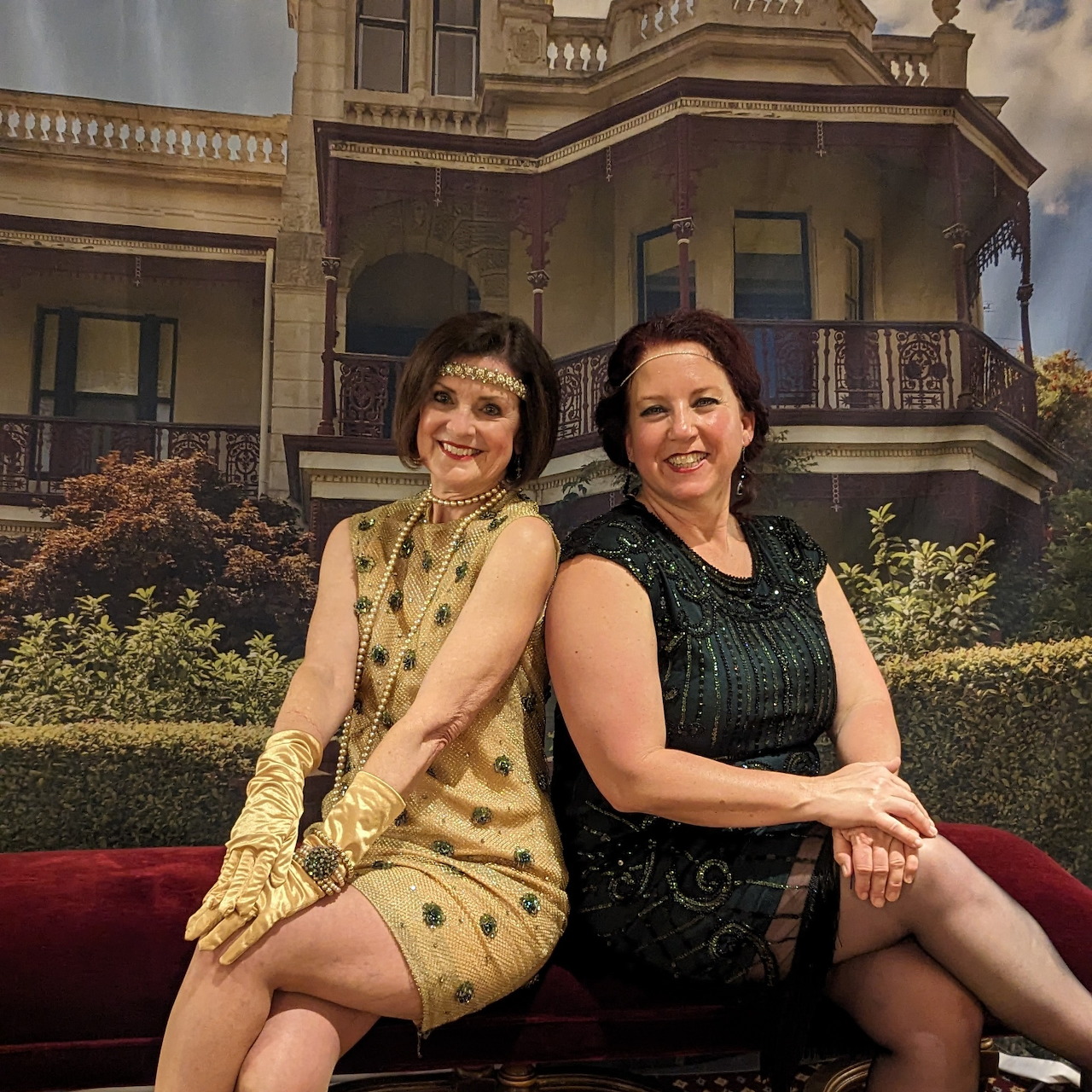 Two women in 1920s dresses sitting on red couch in front of grand Victorian mansion