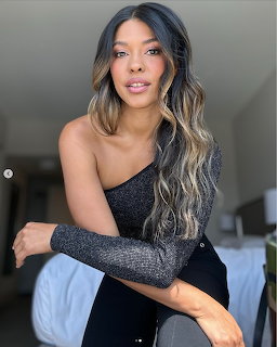 Woman with long wavy brown and blonde hair in a one-shoulder sparkly top posing indoors