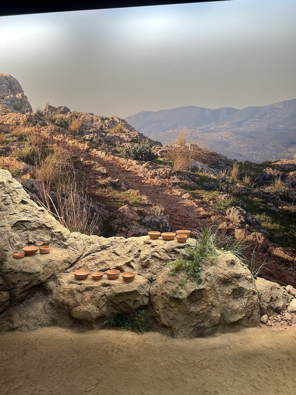 Museum diorama of a rocky desert hillside with small clay pots and distant mountains