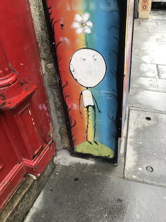 Stick figure with flower painted on rainbow-colored pole next to red door on city sidewalk