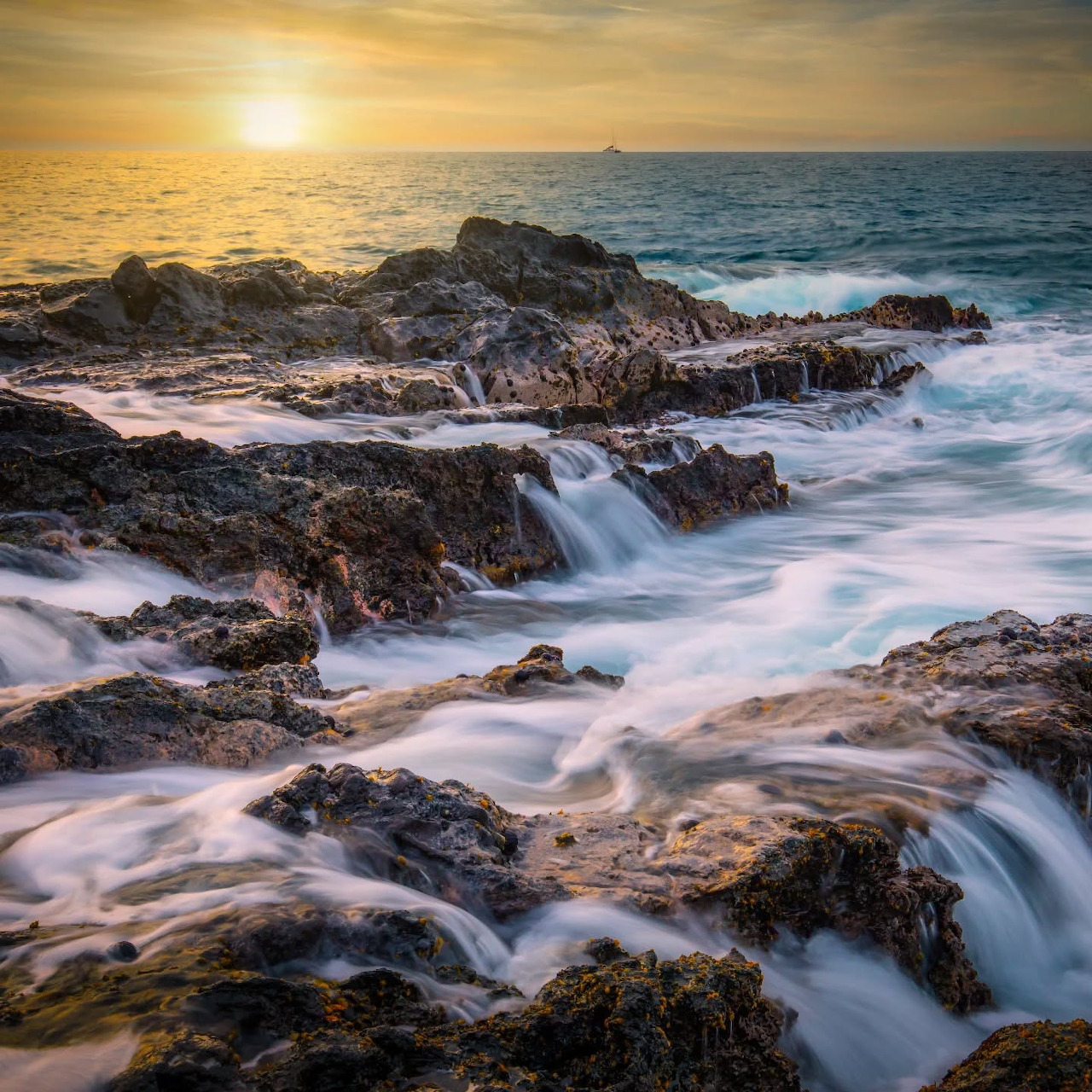 Sunset over a rocky ocean shore with waves and flowing seawater around dark rocks