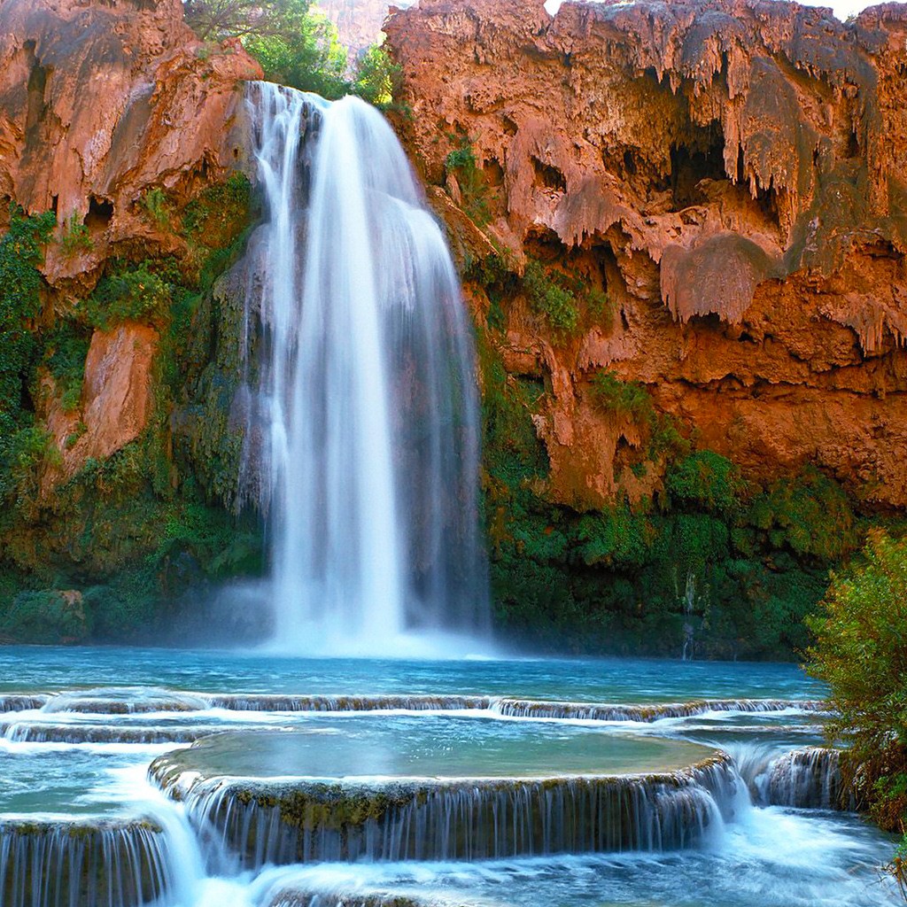 Turquoise waterfall flowing over red rock canyon walls into blue pool with cascading water in foreground