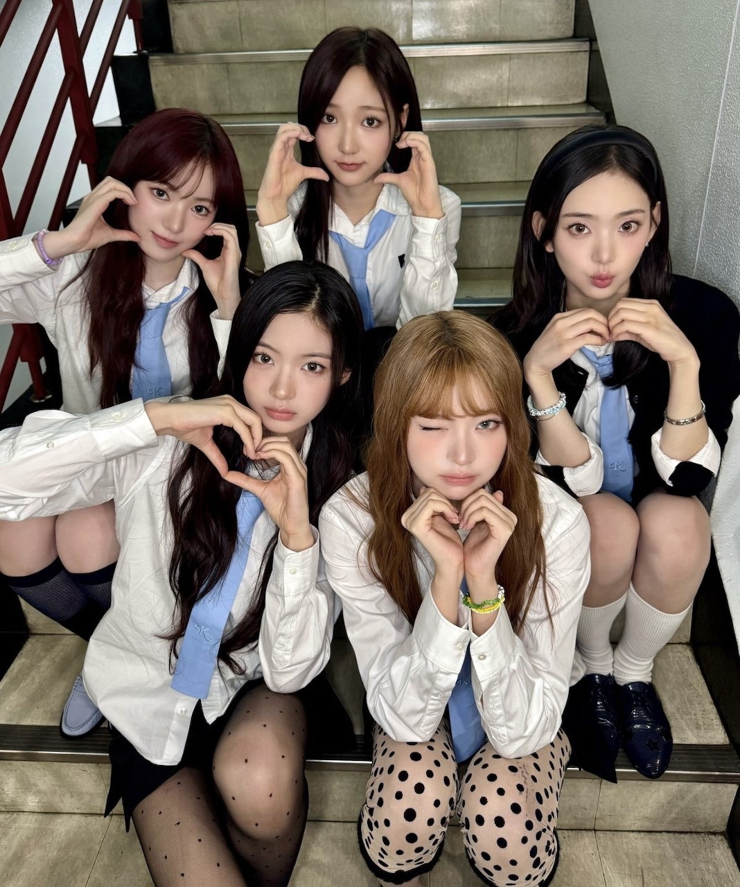 Five K-pop girls in white shirts and blue ties sitting on stairs making heart shapes with their hands