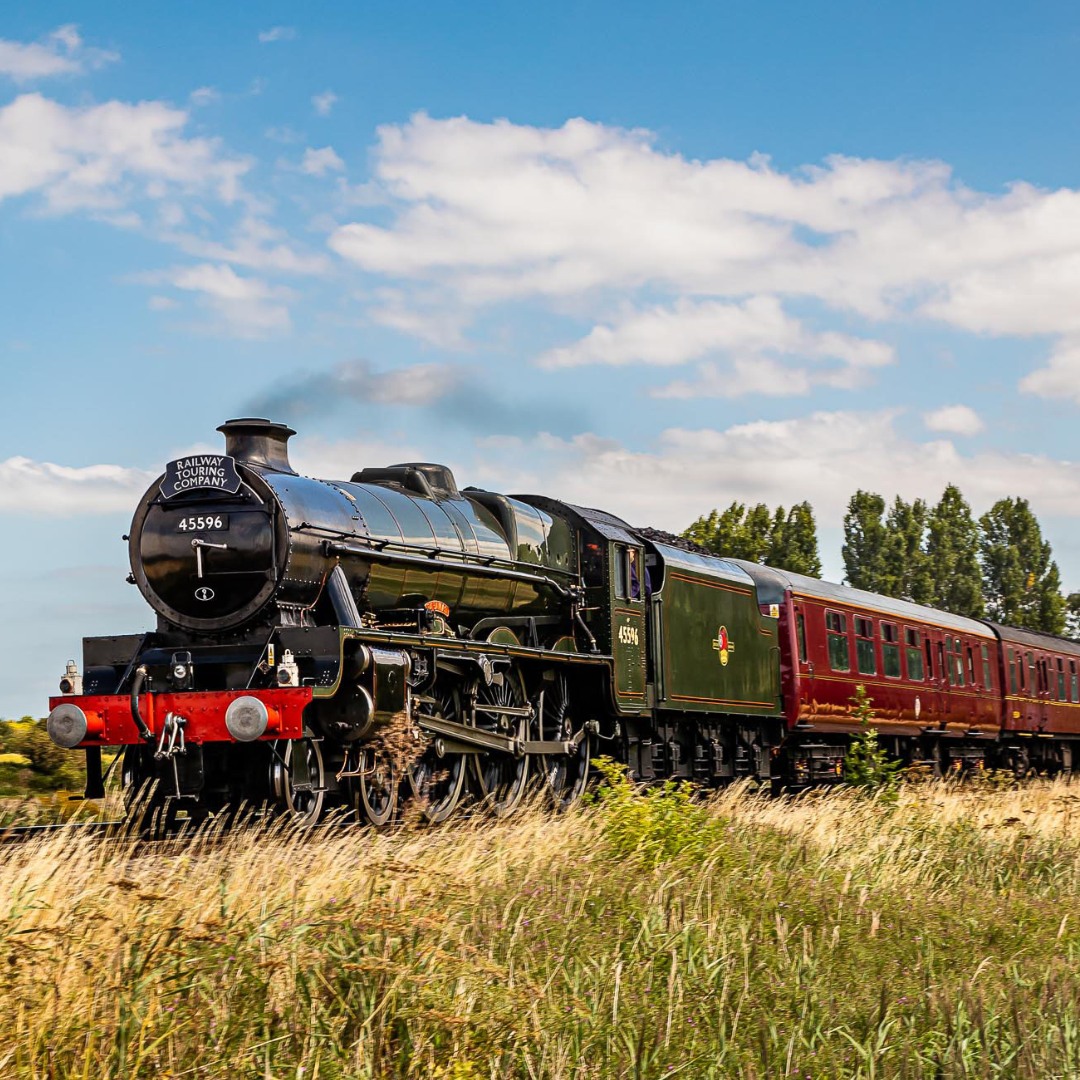 Green vintage steam locomotive with red passenger cars traveling through golden wheat field under cloudy blue sky