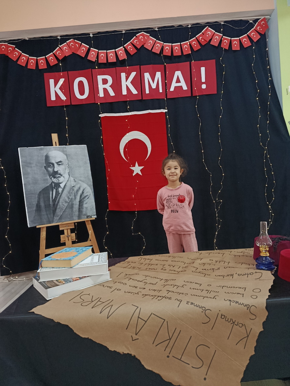 Young girl standing next to an Atatürk portrait on an easel with a Turkish flag and KORKMA banner backdrop