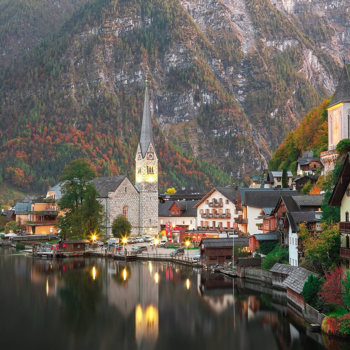 Hallstatt village on lake shore with church tower, mountains behind, autumn colors, dusk reflections in water