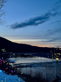 Twilight river view with dark mountain silhouettes, orange sunset glow, and colorful string lights