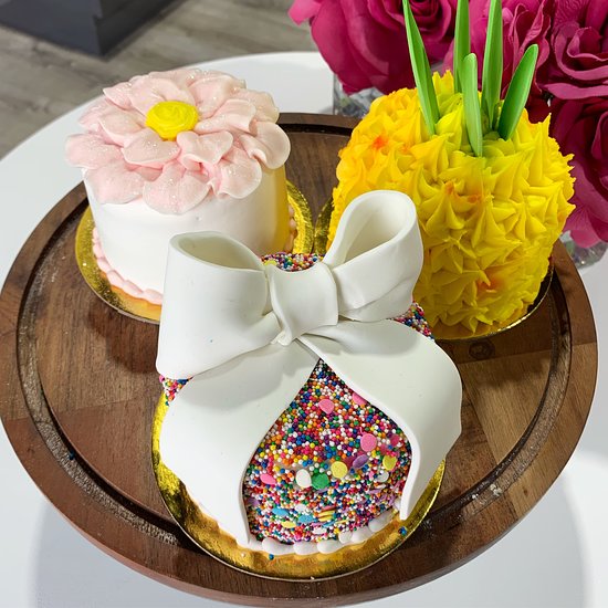 Three decorated mini cakes on a wooden tray with a white bow, flower topper, sprinkles, and yellow frosting