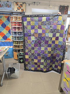 Purple and yellow patchwork quilt hanging on display in a quilting shop with shelves of fabric in background