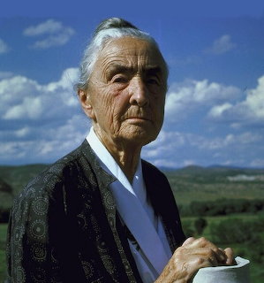 Elderly woman with white hair in dark patterned shawl standing outdoors against blue sky and green hills