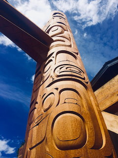 Tall carved wooden totem pole with Indigenous faces and designs against a blue sky with clouds