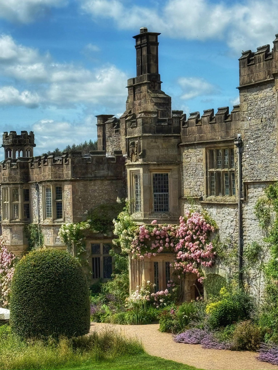 Historic stone manor house with chimneys, towers, and blooming garden shrubs under a blue sky
