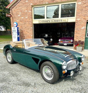 Dark green vintage Healey convertible parked in front of a brick classic car garage