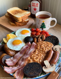 Full English breakfast plate with fried eggs, bacon, sausages, beans, mushrooms, tomatoes, and toast beside a mug of tea