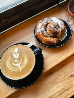 Latte with leaf art in a dark cup next to a glazed cinnamon roll on a wooden table