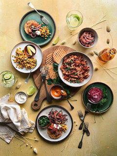 Overhead flatlay of various savory dishes including pulled meat, corn salad, shrimp, and dipping sauces on a golden background.
