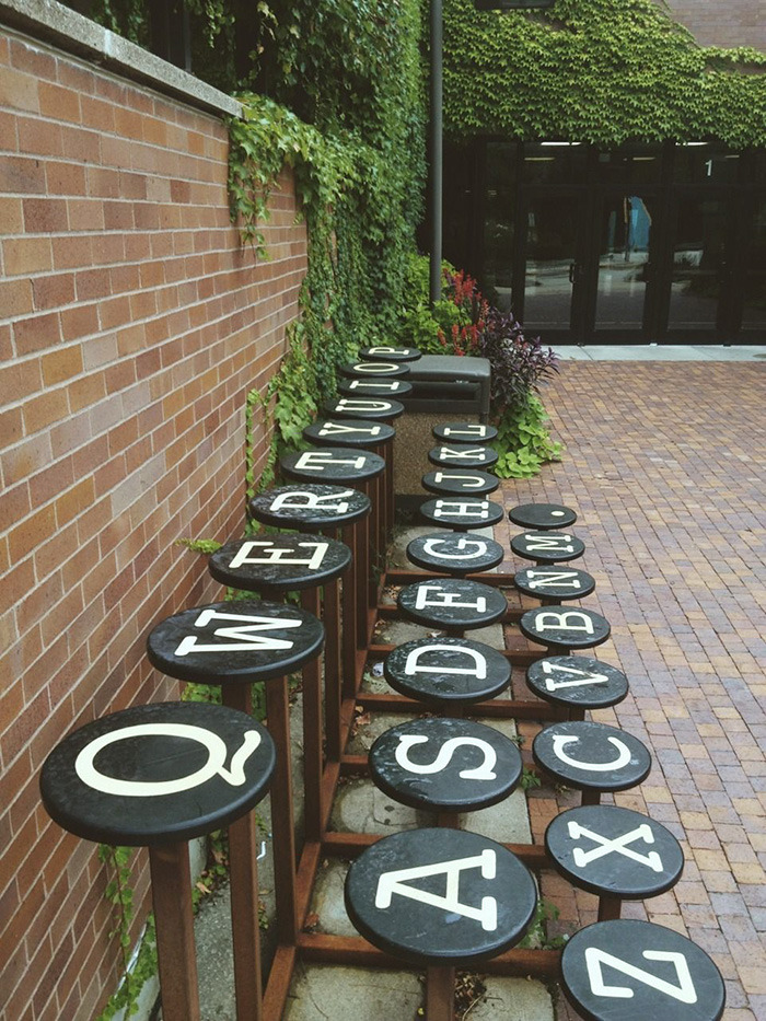 Outdoor bench with round black seats painted as typewriter keys in alphabetical order along a brick path