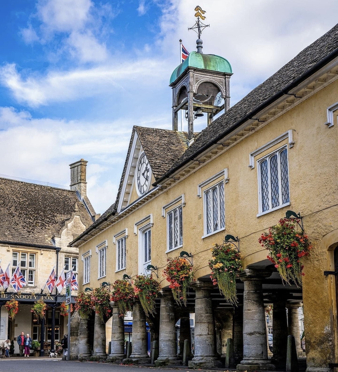 Historic Cotswold town square with yellow stone buildings, clock tower with green cupola, and flower baskets