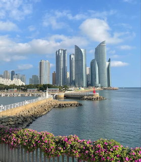 Modern waterfront skyline with tall skyscrapers, calm sea, blue sky, and pink flowers in the foreground.