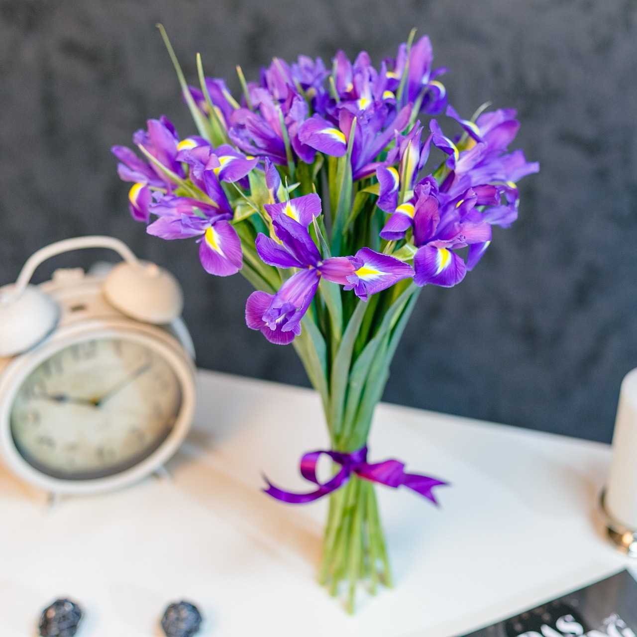 Purple iris bouquet in a vase on a white table beside an alarm clock