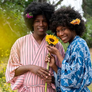 Two women in patterned robes with flowers in their hair holding a sunflower and smiling at camera