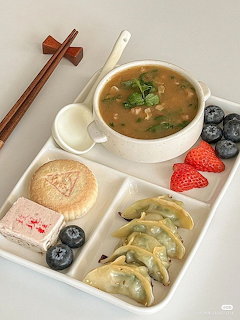 Overhead view of white bento box containing soup, dumplings, rice cake, strawberry, and blueberries with chopsticks
