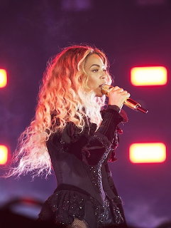 Female singer with blonde curly hair singing into a microphone on stage with red lighting