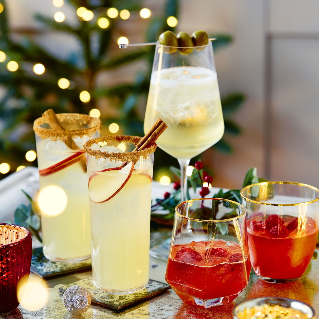 Assorted festive cocktails with fruit garnishes and cinnamon sticks beside Christmas tree lights